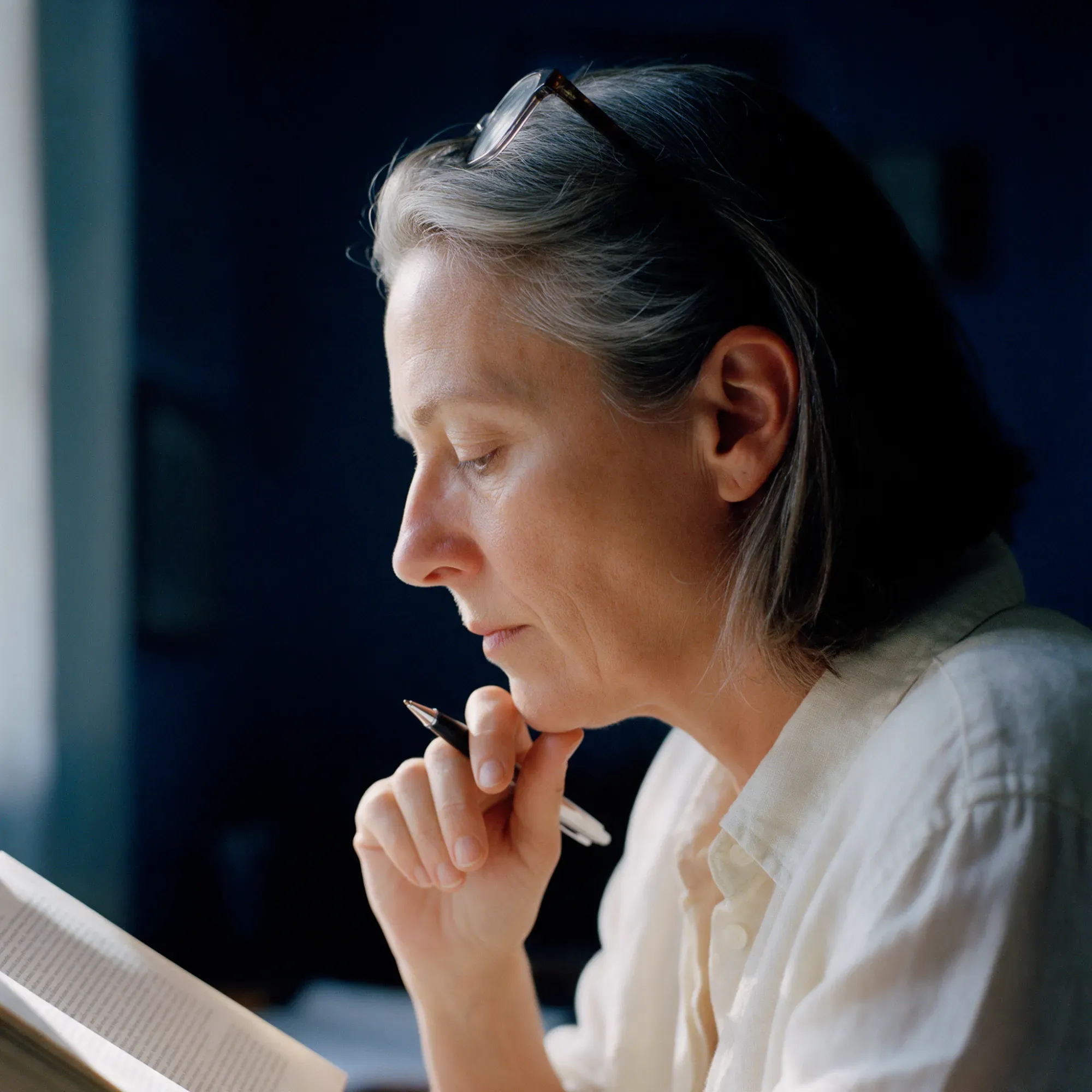 A woman in her mid-fifties in profile, absorbed in reading, a pen held near her chin