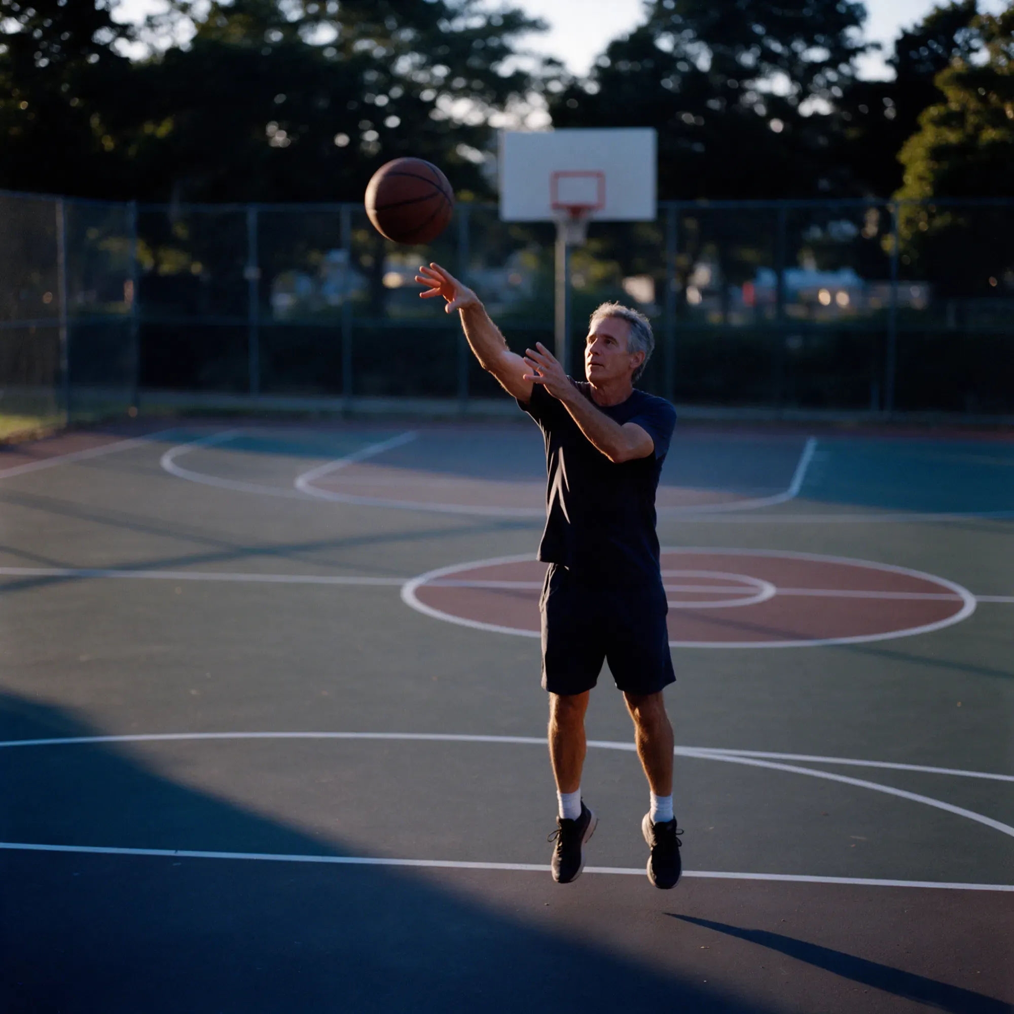 A man in his mid-fifties shooting a basketball on an outdoor court at dusk