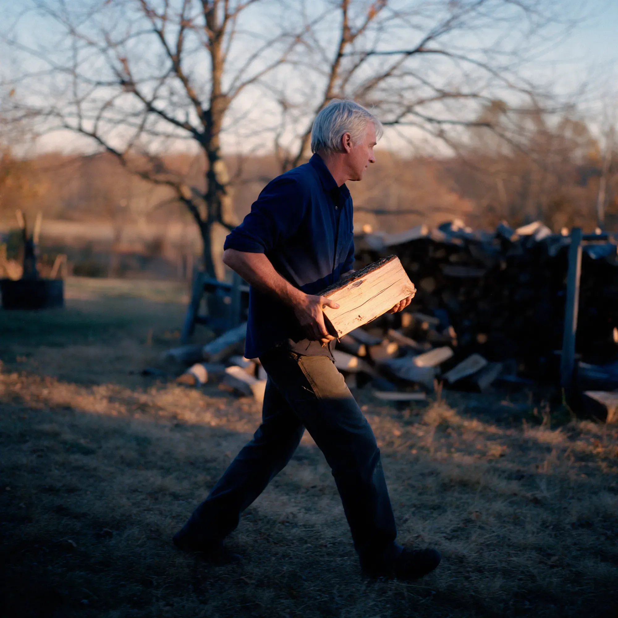 A man in his mid-fifties carrying a split log across a yard at golden hour