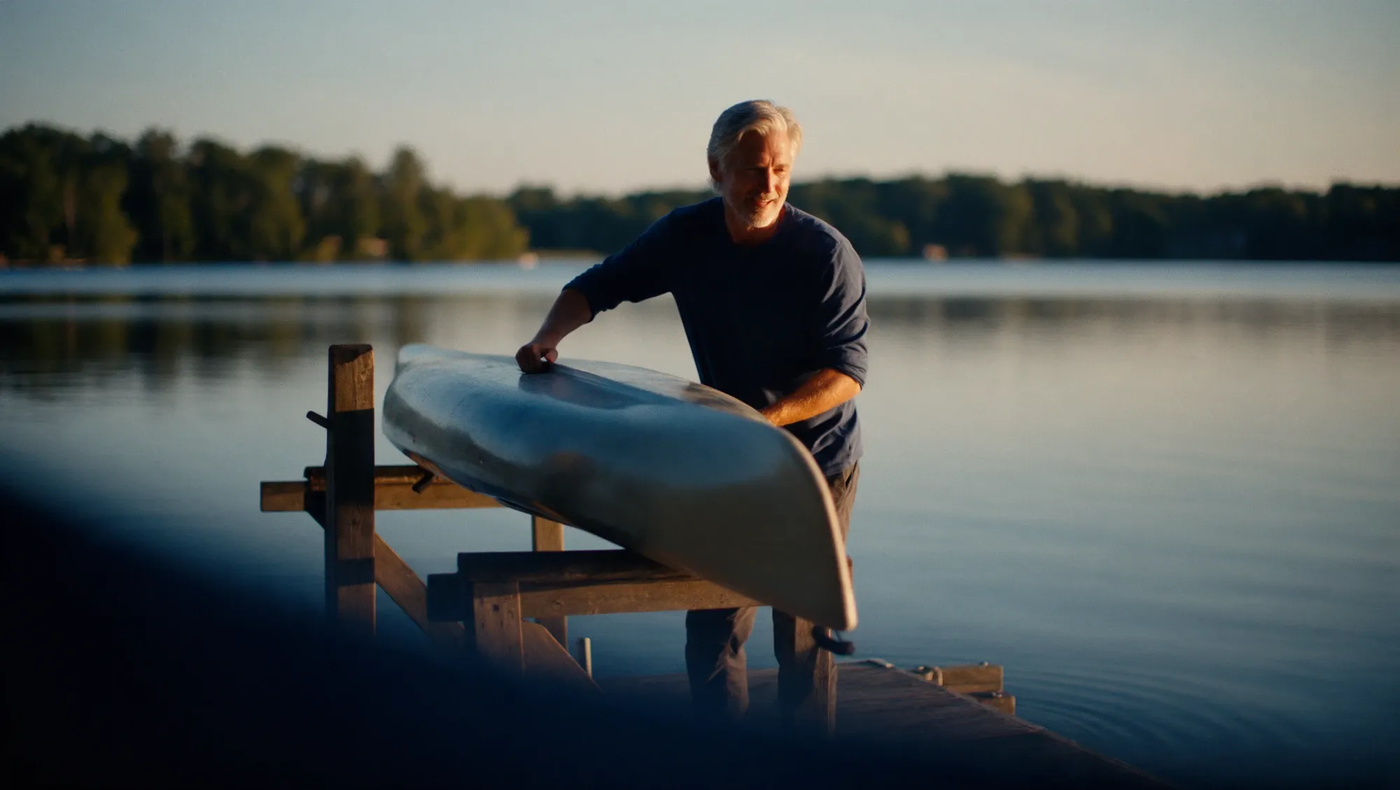 A man in his mid-fifties lifting a kayak onto a wooden rack beside a lake at golden hour