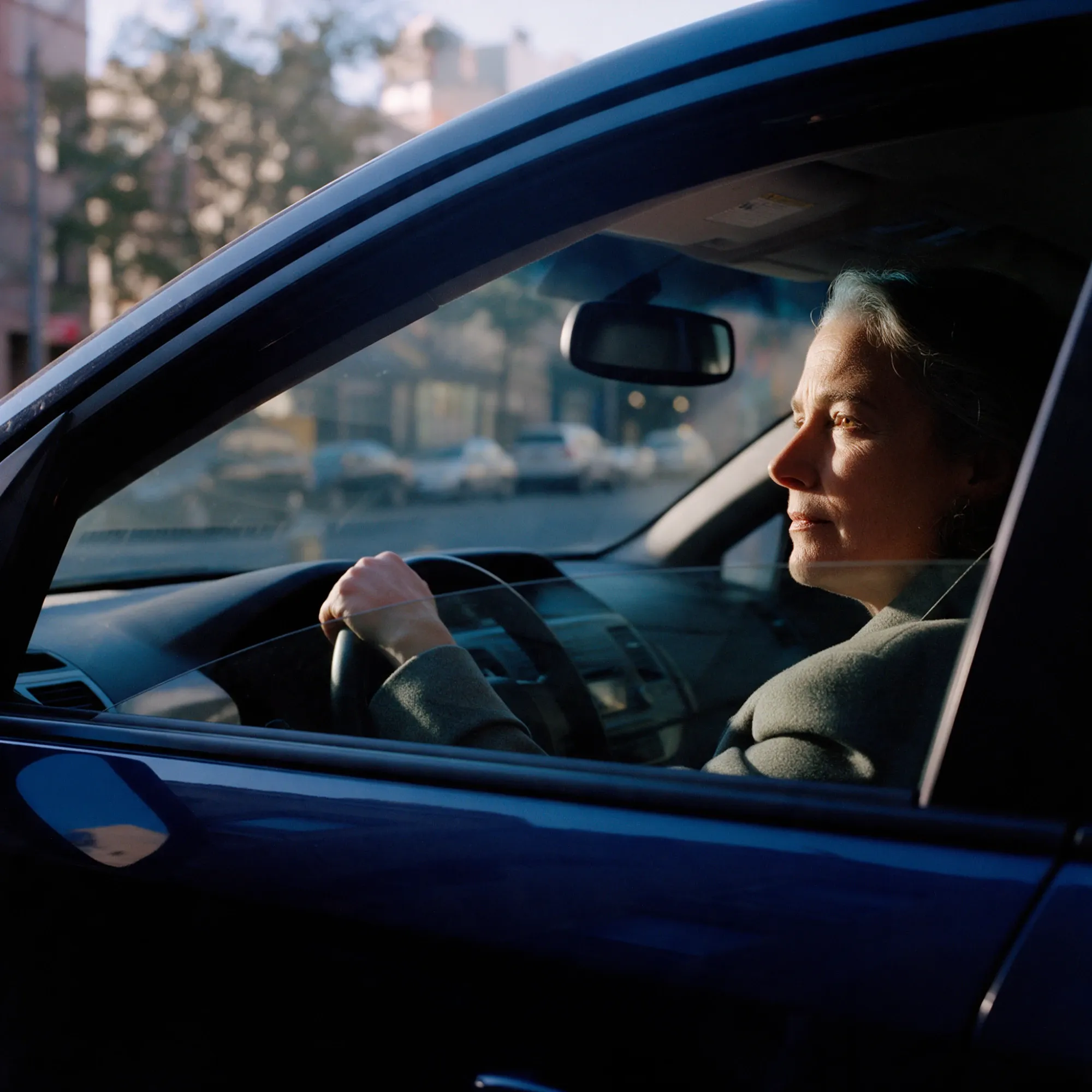 A woman in her mid-fifties in her parked car, hand on the wheel, looking ahead and thinking