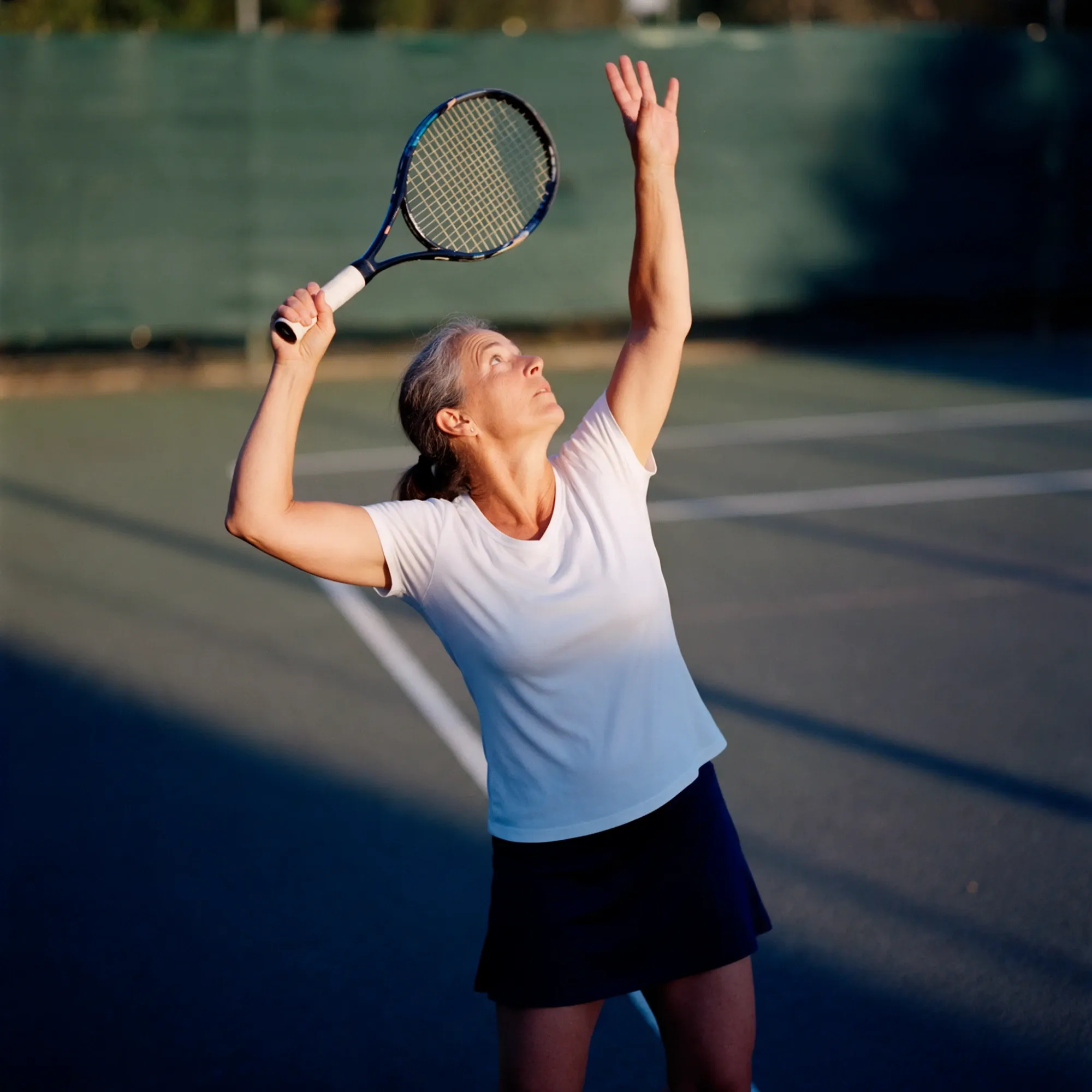 A woman in her mid-fifties serving on an outdoor tennis court at golden hour